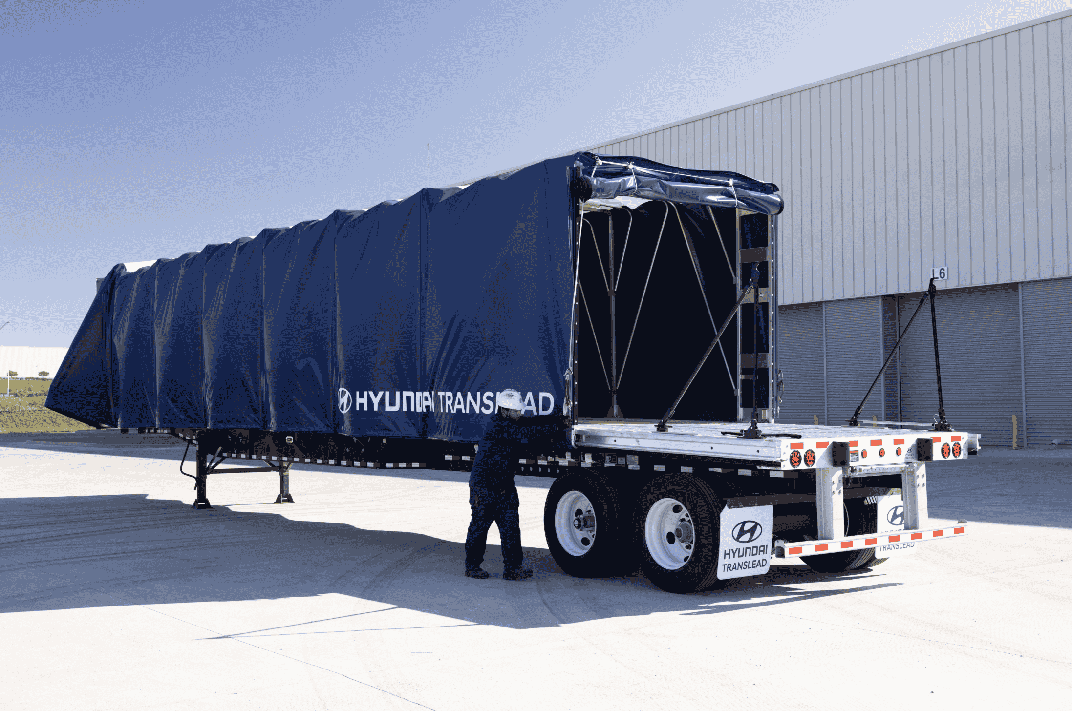 A worker adjusts the blue retractable tarp system on a Hyundai Translead Latitude Flatbed trailer, showcasing Hyundai's innovative trailer technology and design for cargo protection.